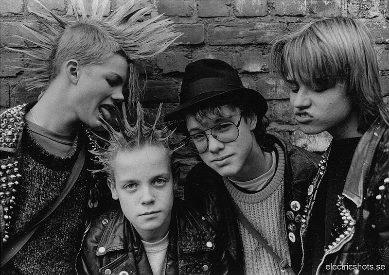 A group of four boys with mowhawks and silly expressions pose together against a brick wall.