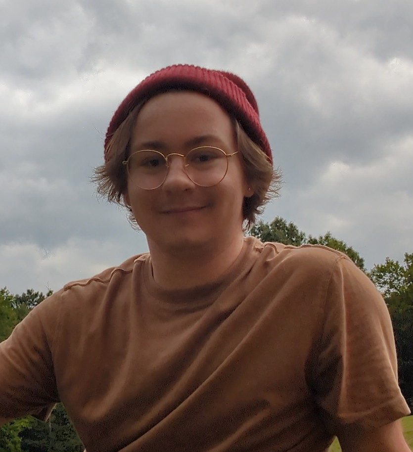 A person wearing glasses, a red beanie, and a brown shirt is outdoors with trees and a cloudy sky in the background.