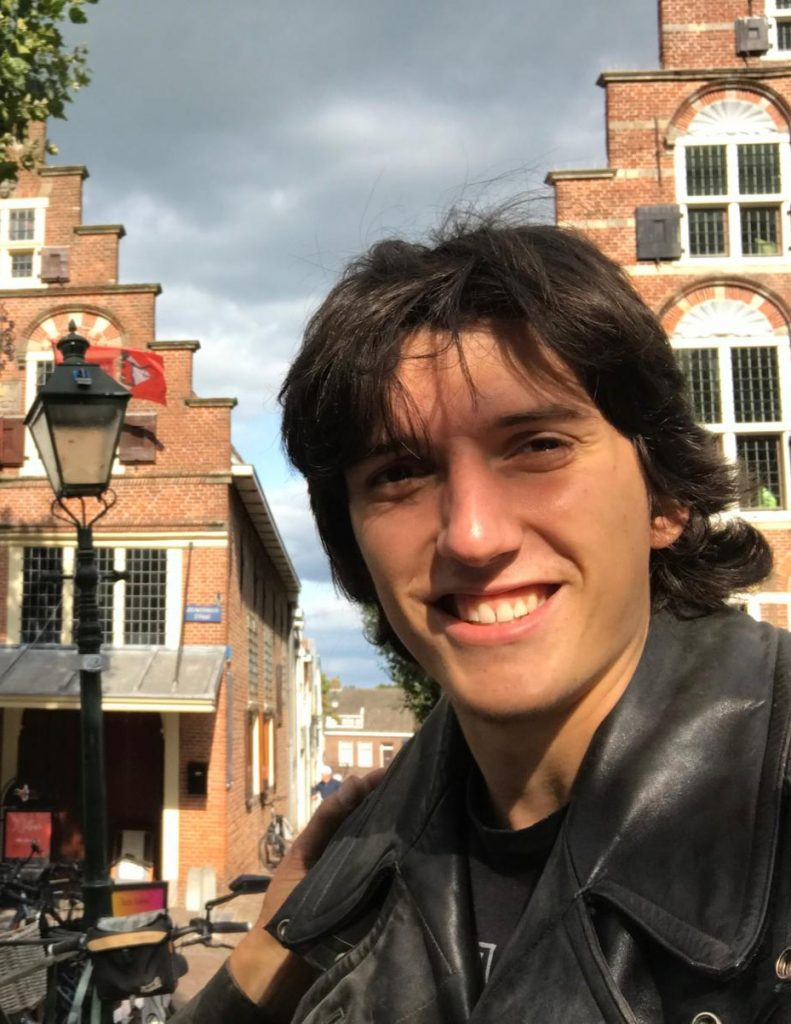 Young person with dark hair in a leather jacket smiling outdoors, with brick buildings and a streetlamp in the background on a cloudy day.