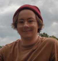 A person wearing glasses, a red beanie, and a brown shirt is outdoors with trees and a cloudy sky in the background.