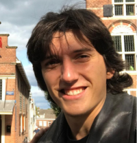 Young person with dark hair in a leather jacket smiling outdoors, with brick buildings and a streetlamp in the background on a cloudy day.